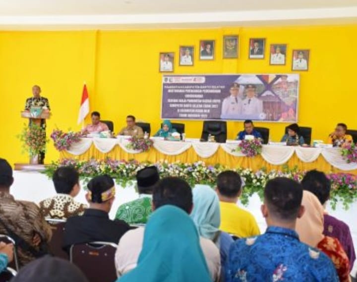 Eddy Raya Samsuri, Bupati Barito Selatan, berdiri di podium pada acara Musrenbang RKPD di ruang rapat berwarna kuning, dengan spanduk dan pejabat lainnya di latar belakang.