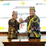 Wakil Gubernur Kalimantan Tengah Edy Pratowo berjabat tangan dengan pejabat lainnya saat menyerahkan LKPD unaudited 2025, keduanya mengenakan batik warna hitam dengan motif khas, latar layar presentasi berlogo.