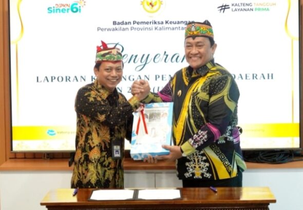 Wakil Gubernur Kalimantan Tengah Edy Pratowo berjabat tangan dengan pejabat lainnya saat menyerahkan LKPD unaudited 2025, keduanya mengenakan batik warna hitam dengan motif khas, latar layar presentasi berlogo.