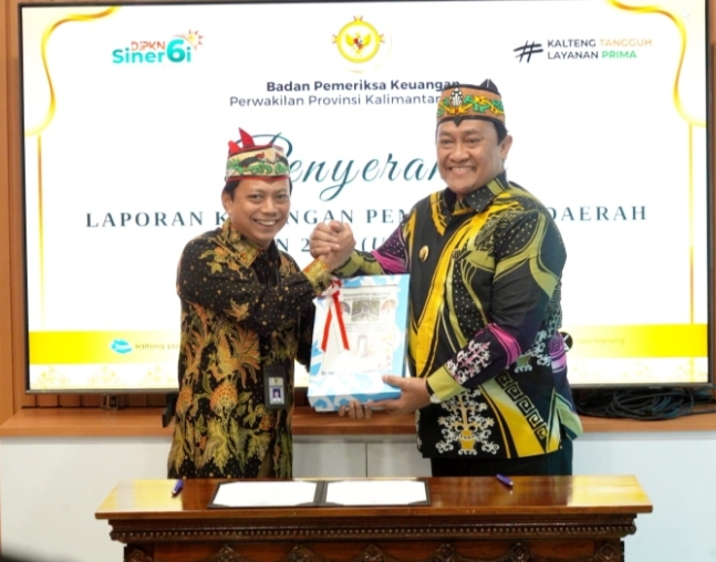 Wakil Gubernur Kalimantan Tengah Edy Pratowo berjabat tangan dengan pejabat lainnya saat menyerahkan LKPD unaudited 2025, keduanya mengenakan batik warna hitam dengan motif khas, latar layar presentasi berlogo.