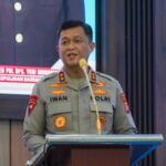 Irjen Pol Iwan Kurniawan berbicara di podium saat Bakti Kesehatan Bhayangkara ke-80 di Palangka Raya, dengan layar presentasi di belakangnya.