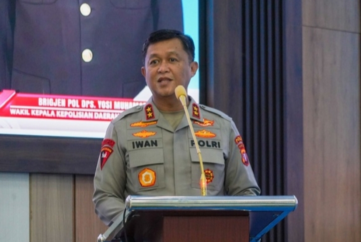 Irjen Pol Iwan Kurniawan berbicara di podium saat Bakti Kesehatan Bhayangkara ke-80 di Palangka Raya, dengan layar presentasi di belakangnya.